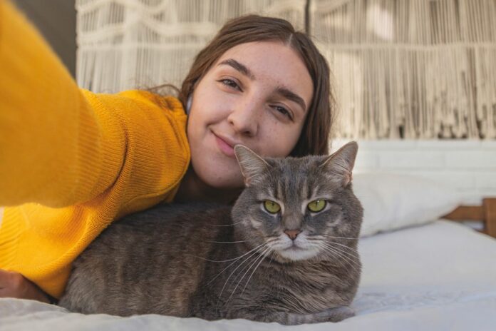 Une femme souriante prenant un selfie avec un chat posé sur un lit.